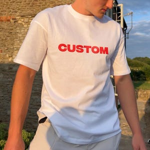 May include: A white short-sleeved t-shirt featuring the word CUSTOM printed in bold red capital letters across the centre of the chest. The garment has a classic crew neckline and a relaxed fit, constructed from a smooth, solid white fabric.