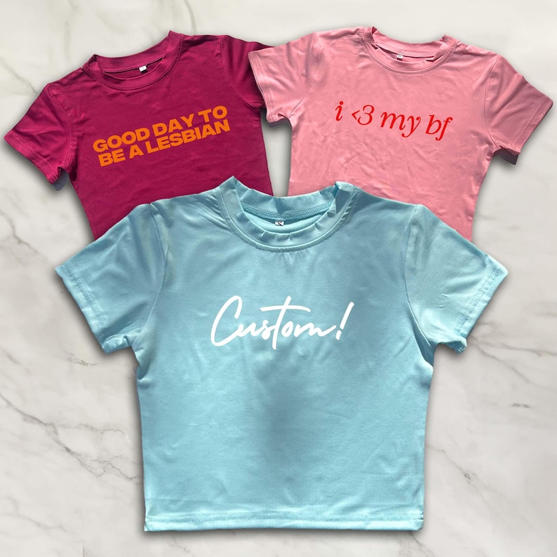 Puede incluir: Tres camisetas recortadas con diferentes lemas. La primera es una camiseta granate con el texto "Good day to be a lesbian" en naranja. La segunda es una camiseta rosa claro con el texto "i <3 my bf" en rojo. La tercera es una camiseta azul claro con el texto "Custom!" en blanco.