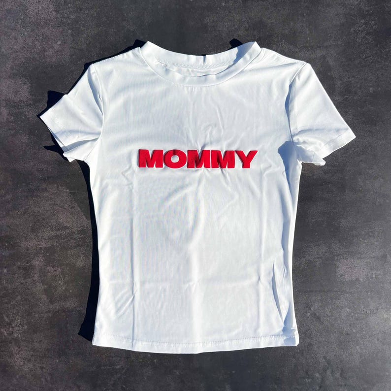 May include: White short-sleeved t-shirt with the word "MOMMY" in red raised letters.