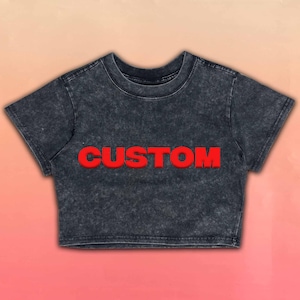 May include: A dark gray crop top with the word "CUSTOM" in bold red letters across the front. The shirt has a crew neck and short sleeves. The fabric has a textured, stone-washed appearance.