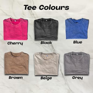 May include: Six t-shirts in various colors are displayed, including cherry, black, blue, brown, beige, and grey. The text "Tee Colours" is at the top. Each shirt is folded and presented on a marble-patterned surface.