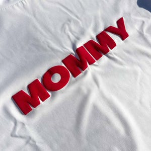 May include: White t-shirt with the word "MOMMY" in large, red, raised letters.