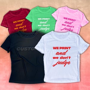 Puede incluir: Cinco camisetas de manga corta en varios colores: burdeos, verde, rosa, negro y blanco. Cada camiseta presenta la frase "WE PRINT and we don't judge" en colores contrastantes. La camiseta negra tiene la palabra "CUSTOM" impresa.