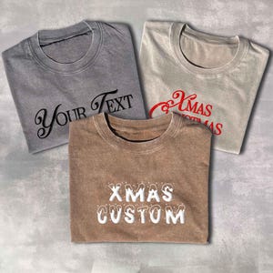 Può includere: Tre magliette girocollo grigie, beige e marroni. La maglietta grigia ha "Your Text" in corsivo nero. La maglietta beige ha "Xmas" in corsivo rosso. La maglietta marrone ha "XMAS CUSTOM" in bianco.