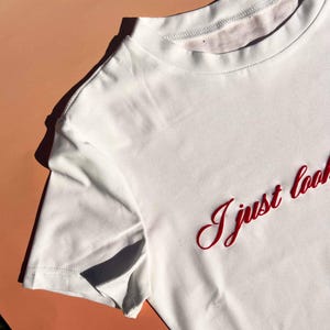 May include: White t-shirt with red embroidered text that says "I just look"