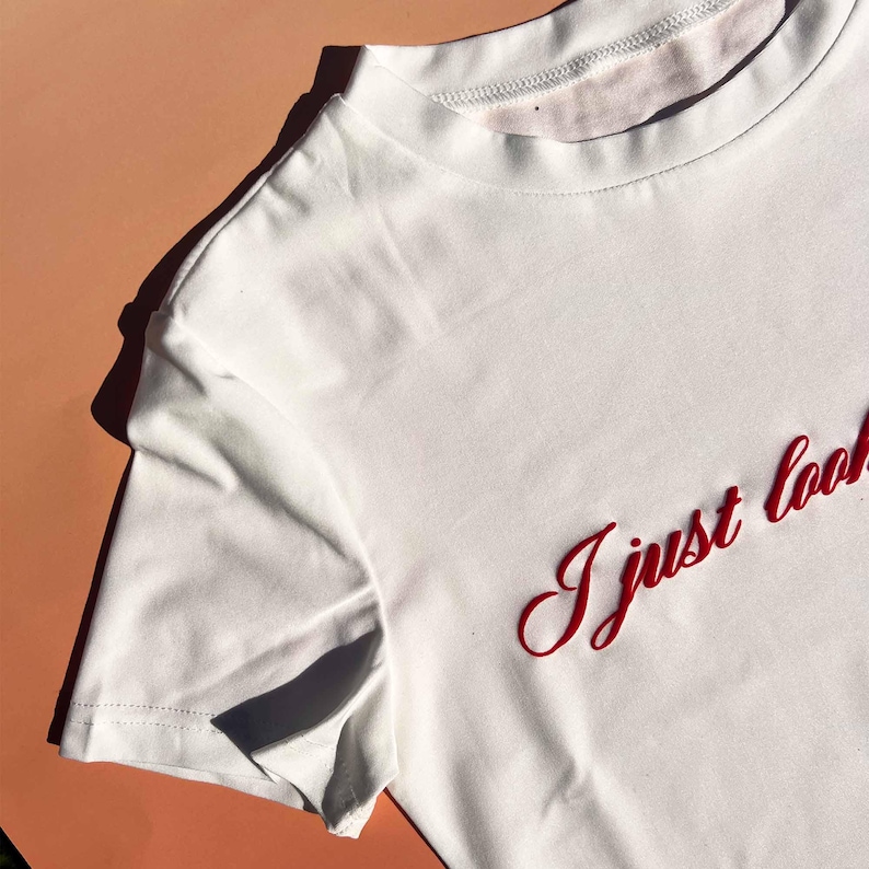 May include: White t-shirt with red embroidered text that says "I just look"