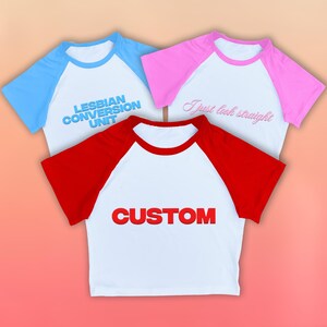 May include: Three baseball-style shirts with white bodies and colored sleeves. One shirt has blue sleeves and the text "LESBIAN CONVERSION UNIT". Another has pink sleeves and the text "I just look straight". The third has red sleeves and the text "CUSTOM".