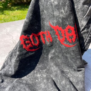 May include: A dark gray t-shirt with a distressed, textured appearance. The shirt features the word "GOTH DA" in a bold, red, stylized font across the front. The fabric has a mottled, faded effect.