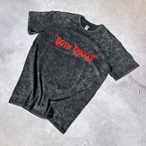 May include: A dark gray, acid-wash t-shirt with the red text "GOTH DADDY" in a stylized font. The short-sleeved shirt has a crew neck and a slightly faded appearance, suggesting a vintage or worn look.
