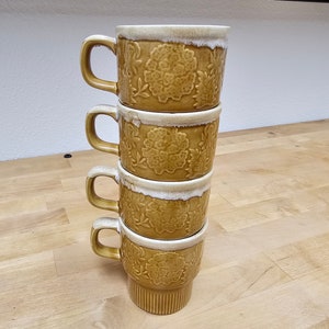 May include: Four stacked ceramic mugs with a floral design in shades of brown and white. The mugs have a white rim and a slightly flared lip.
