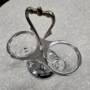 May include: A silver-colored condiment holder with two clear glass cups. The holder has a heart-shaped handle with gold-colored accents. The base is round and reflective.