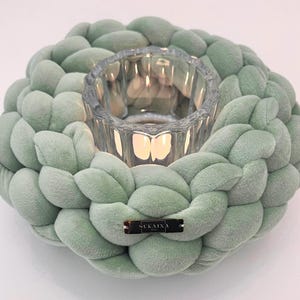 Peut inclure: Un bougeoir en velours tressé vert sauge avec un photophore en verre transparent au centre. Le bougeoir porte une petite étiquette métallique rectangulaire avec l'inscription "SUKAINA". Une bougie allumée est à l'intérieur.