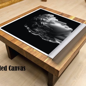 May include: A black and white rolled canvas print of a classical sculpture of a man's head. The canvas is rolled and sitting on a wooden table.