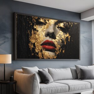 May include: A framed artwork depicting a woman's face with closed eyes and red lips, adorned with gold leaf. The background is black, and the frame is grey. The artwork is displayed above a white sofa.