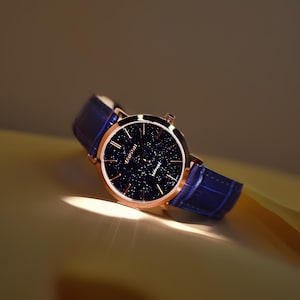 May include: A rose gold watch with a blue leather strap and a dark blue face with a starry pattern. The watch face says "Kienici" and "Sapphire".