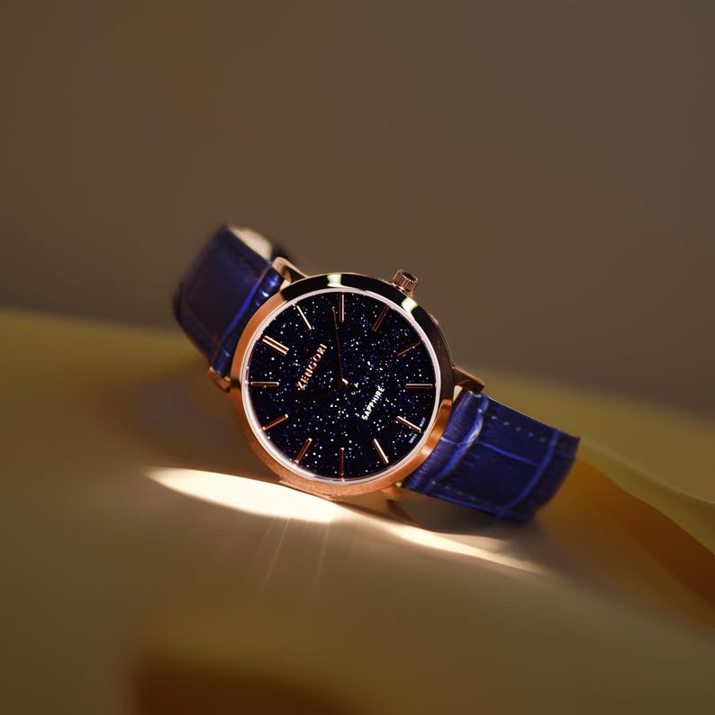 May include: A rose gold watch with a blue leather strap and a dark blue face with a starry pattern. The watch face says "Kienici" and "Sapphire".