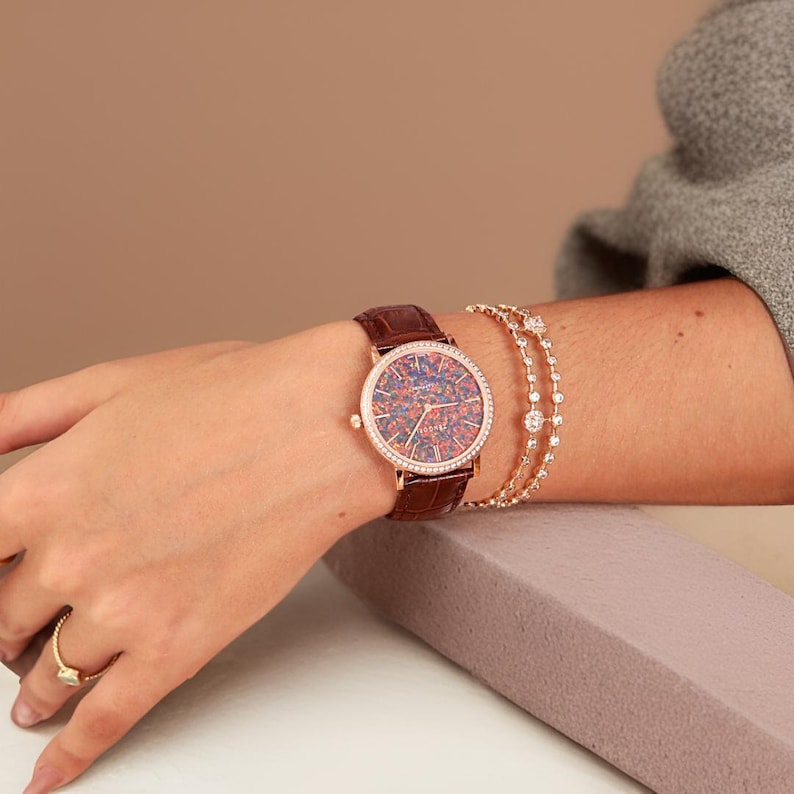 Puede incluir: Un reloj de oro rosa con una correa de cuero marr&oacute;n y una esfera de &oacute;palo de colores. El reloj est&aacute; engastado con diamantes alrededor del bisel.