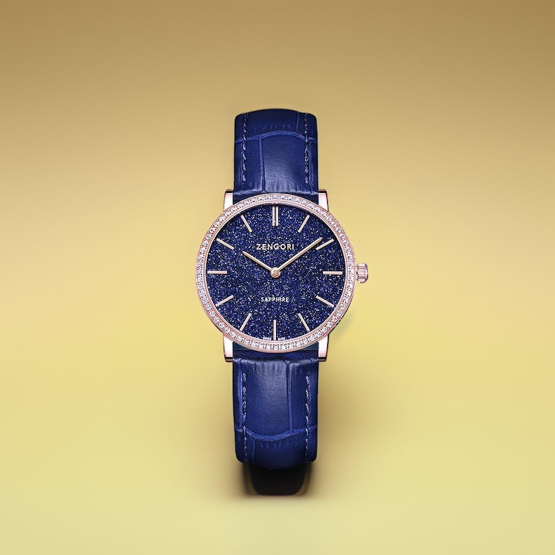 May include: A blue leather wristwatch with a rose gold case and a blue dial with a starry pattern. The watch face has a diamond bezel and the text "ZENGORI SAPPHIRE" printed on it.