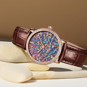 Puede incluir: Un reloj de oro rosa con una correa de cuero marr&oacute;n y una esfera redonda. La esfera est&aacute; cubierta de &oacute;palo iridiscente y rodeada por una fila de cristales transparentes.