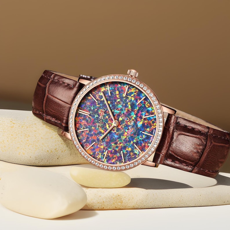 Puede incluir: Un reloj de oro rosa con una correa de cuero marr&oacute;n y una esfera redonda. La esfera est&aacute; cubierta de &oacute;palo iridiscente y rodeada por una fila de cristales transparentes.