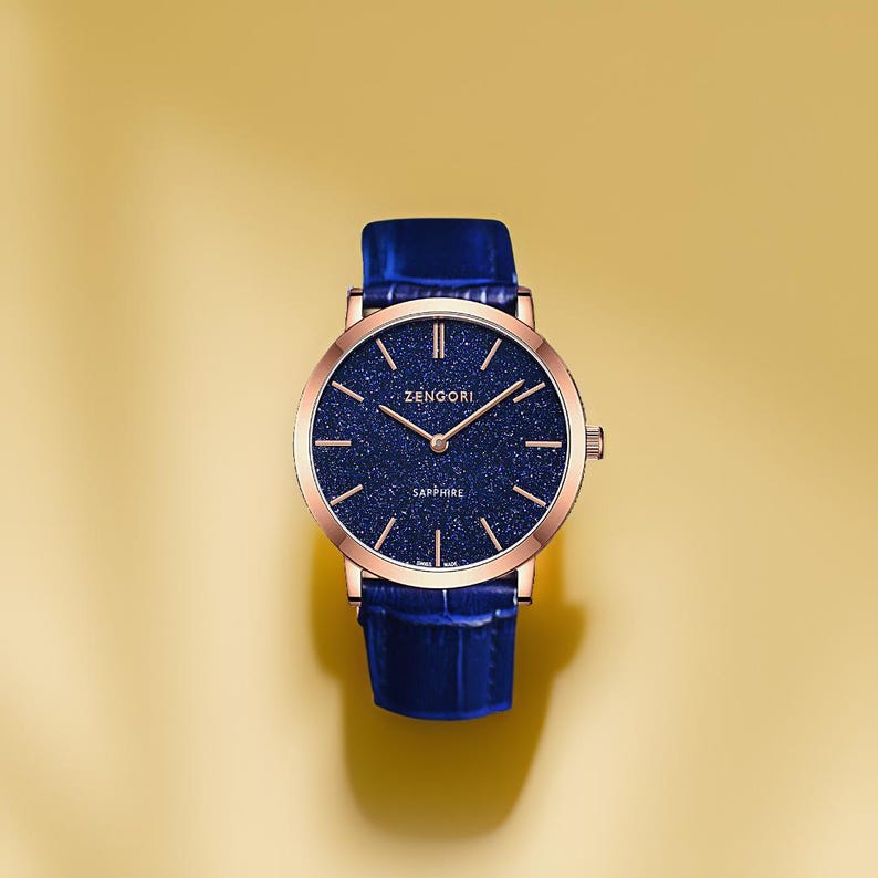 May include: A rose gold watch with a blue leather strap and a blue face with a sparkly texture. The face of the watch has the words "ZENGORI" and "SAPPHIRE" printed on it.