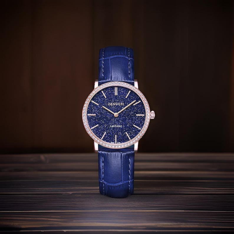 May include: A blue leather wristwatch with a rose gold case and a blue starry dial. The watch face has a diamond bezel and the text "ZENGORI SAPPHIRE" printed on it.