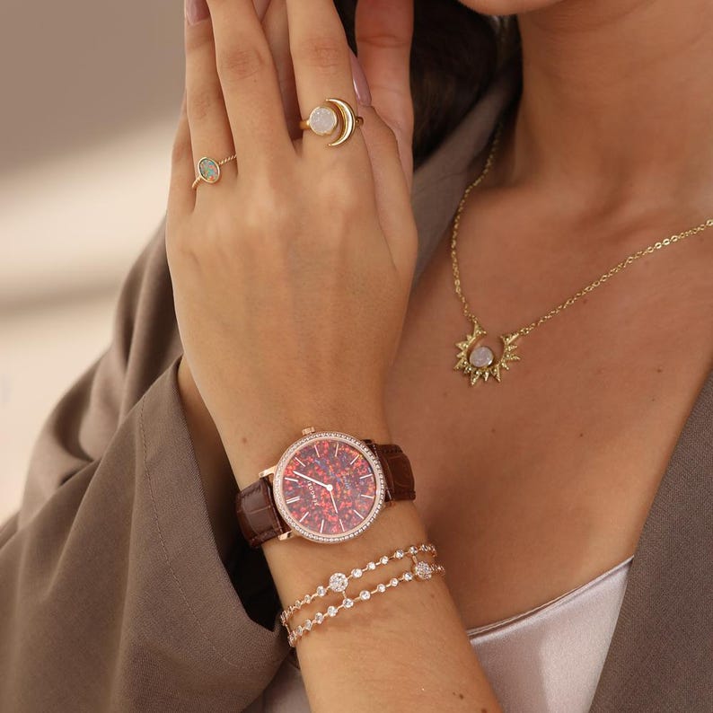 Puede incluir: La mu&ntilde;eca de una mujer con un reloj de cuero marr&oacute;n con una esfera roja y detalles de diamantes. El reloj tiene una correa dorada y se combina con una pulsera de diamantes delicada. La mujer tambi&eacute;n lleva un collar de oro con un colgante de sol y luna.