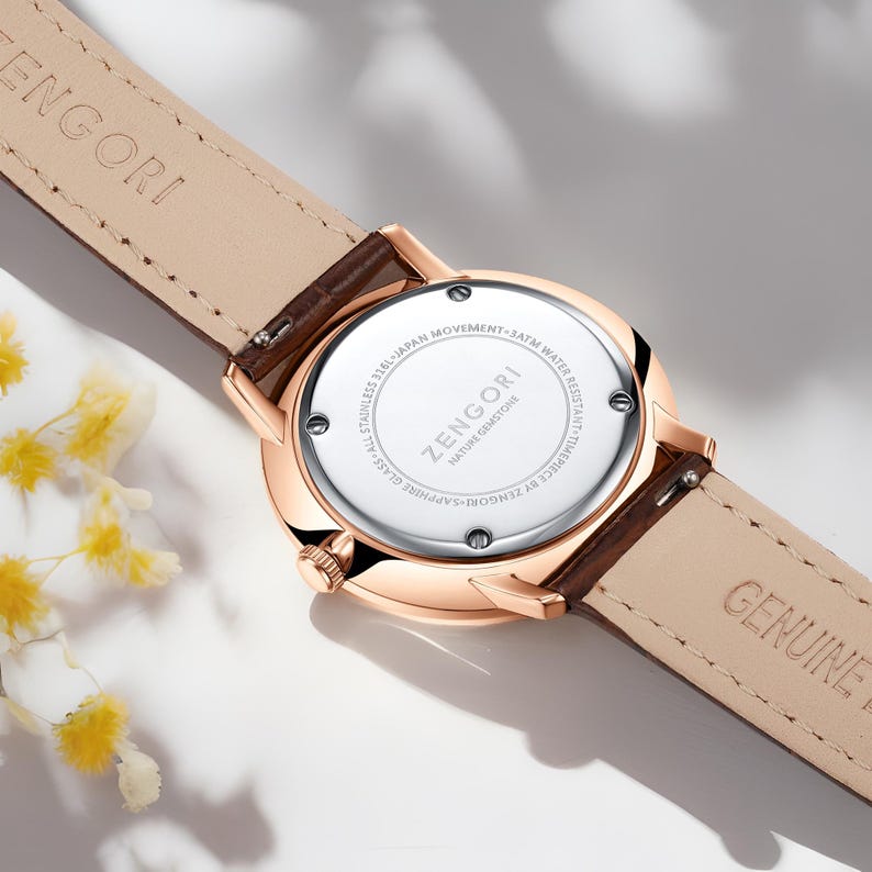 Puede incluir: Un reloj de oro rosa con una correa de cuero marr&oacute;n. La parte posterior del reloj tiene el texto "ZENGORI" y "NATURE GEMSTONE" impreso. El reloj es resistente al agua hasta 3 ATM.