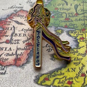 Puede incluir: Un pin de color dorado con una pancarta blanca que dice "FOLLANDS" en letras azules. La pancarta está decorada con una cinta morada, verde y amarilla. El pin está colocado sobre un mapa antiguo de las Islas Británicas.