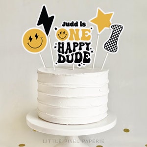 Könnte beinhalten: Ein weißer Kuchen mit gelben und schwarzen Kuchentoppern. Die Topper sagen "Judd is One Happy Dude" und zeigen ein Smiley, einen Stern, einen Blitz und eine karierte Flagge.