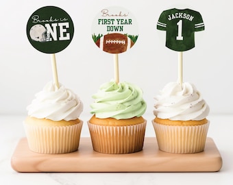 May include: Three cupcakes decorated with green, white, and brown frosting. Each cupcake has a different football-themed topper with the text "Brooks is One", "Brooks' First Year Down", and "Jackson 1".