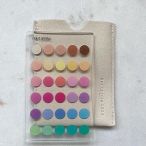 May include: A clear rectangular acrylic card featuring the text LIGHT SPRING and a grid of thirty circular color swatches in various shades. The card is placed on a beige fabric sleeve with the text rose and taupe printed vertically.