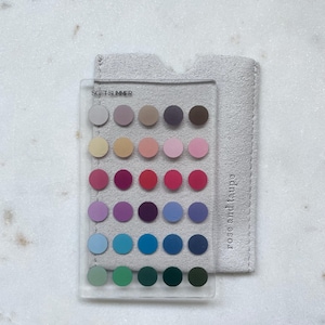 May include: A clear rectangular acrylic color palette card featuring the text SOFT SUMMER and thirty small circular color swatches in various shades, layered over a light gray textured fabric sleeve with the text rose and taupe printed on it.