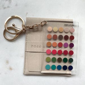 May include: A clear acrylic keychain featuring a grid of thirty small colored circles, labeled "DARK AUTUMN" and "rose and taupe". The keychain includes a gold-toned metal ring and chain, resting on a light beige fabric pouch.