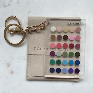 May include: A clear rectangular acrylic keychain featuring a grid of thirty small colored circles and the text SOFT AUTUMN and rose and taupe. The keychain is attached to a gold-toned metal split ring and chain, resting on a beige fabric pouch.