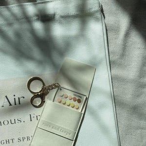 Może przedstawiać: Biała pł&oacute;cienna torba z czarnym napisem, zestawiona z małym, jasnym prostokątnym etui. Etui posiada złoty brelok i zawiera przezroczystą akrylową kartę z siatką małych, okrągłych pr&oacute;bek kolor&oacute;w w r&oacute;żnych odcieniach.