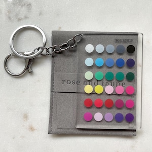 May include: A clear acrylic keychain featuring a grid of thirty colored circles arranged in six rows of five. The text TRUE WINTER is printed at the top, and rose and taupe is printed in the center. It includes a silver metal split ring.