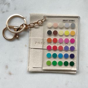 May include: A clear rectangular acrylic keychain featuring a grid of thirty small colored circles. The text BRIGHT SPRING is printed at the top, and rose and taupe is embossed on the beige fabric pouch underneath the acrylic piece.