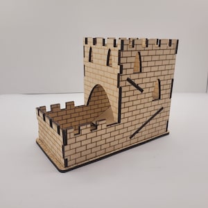 May include: A wooden castle-shaped dice tower with a brick pattern and a cutout archway. The tower is made of light brown wood with black lines outlining the bricks.
