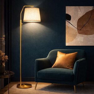 May include: A gold floor lamp with a beige lampshade illuminates a teal armchair with an orange pillow. The lamp has a circular base and a curved arm. A painting with abstract shapes hangs on the dark blue wall.
