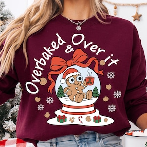May include: A maroon crewneck sweatshirt featuring a graphic of a gingerbread person inside a snow globe. The text reads Overbaked & Over it. The design includes a red bow, green trees, a mug, presents, a candy cane, and white snowflakes.
