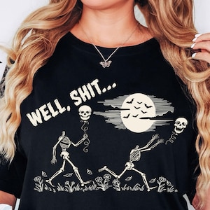 May include: A black t-shirt featuring a graphic print of two skeletons in a field with flowers. One skeleton holds a skull-shaped balloon, while the other reaches for a floating skull balloon. A full moon with bats and the text "WELL, SHIT..." appear above.