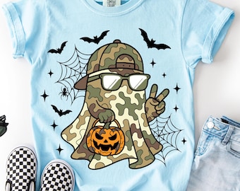 May include: A light blue t-shirt featuring a graphic of a ghost wearing a camouflage baseball cap and sunglasses, making a peace sign while holding a jack-o'-lantern. The design includes black spiderwebs, bats, and stars surrounding the ghost.