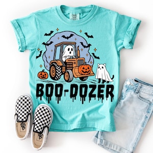 Pode incluir: Uma camiseta turquesa com um gráfico de Halloween. O design apresenta um fantasma dirigindo um trator laranja com uma abóbora. O texto "BOO-DOZER" está abaixo do trator, com gotejamento preto. Sapatos slip-on xadrez preto e branco estão ao lado.