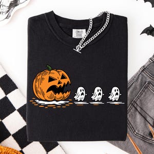 Puede incluir: Sudadera negra con un diseño de Halloween. El diseño presenta una calabaza naranja con una cara tallada y tres figuras de fantasmas blancos. Una cadena plateada está sobre la parte superior. La marca "Comfort Colors" está impresa.
