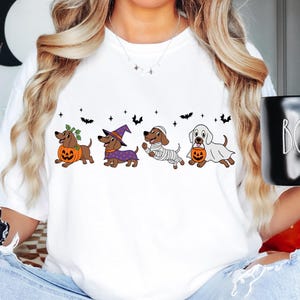 Puede incluir: Una camiseta blanca con un gráfico de cuatro perros salchicha marrones vestidos con disfraces de Halloween. Los perros aparecen como calabaza, bruja, momia y fantasma, rodeados de pequeños murciélagos negros y estrellas impresas en el pecho.