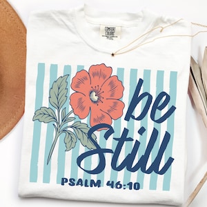 以下が含まれることがあります： 白いTシャツ。紺色の「be Still」の文字と、サンゴ色の花と水色のストライプのフローラルデザイン。 「詩篇46:10」のテキストもプリントされています。