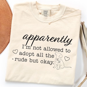 Può includere: T-shirt color crema con la scritta nera "apparently I'm not allowed to adopt all the - rude but okay." Presenta una grafica di un cane che abbraccia un altro cane e due cuori.