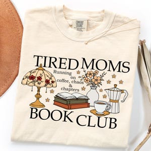 Op de afbeelding: Crèmekleurig T-shirt met de tekst "TIRED MOMS BOOK CLUB" en illustraties van een lamp, bloemen, boeken, koffie en een koffiepot. Het shirt heeft ook de tekst "Running on coffee, chaos & chapters."