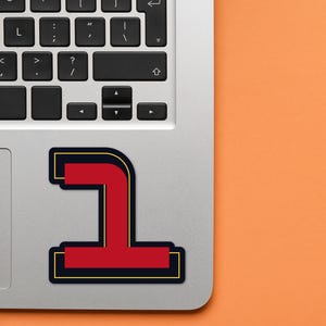 May include: A red and black number one sticker on a silver laptop. The number one is outlined in black and has a gold outline.